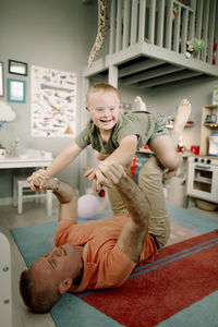 Full length of father lying on carpet while lifting son in bedroom at home