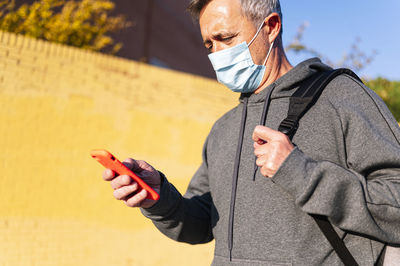 Senior retired man with face mask, sportswear and smart phone walking down the street