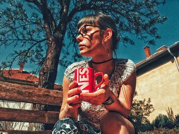 Portrait of young woman holding sunglasses against trees