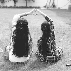Rear view of friends with long hair making heart shape while sitting on land