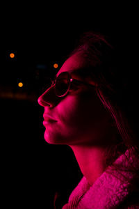 Crop young female in stylish sunglasses in dark studio with pink neon illumination and looking away with gentle smile
