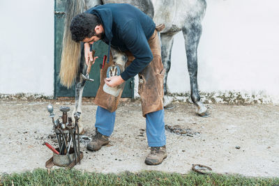 Man in dirty chaps fixing hoof from horse during work in yard outside stable on ranch