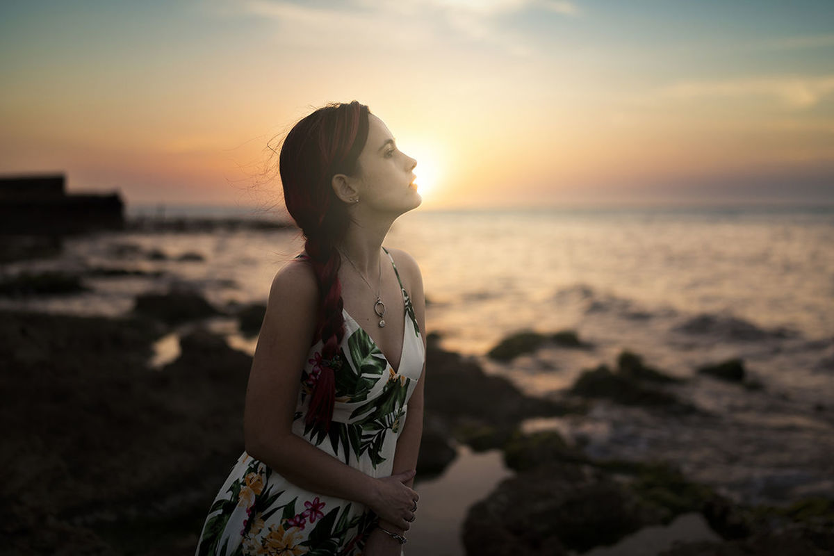 sky, sunset, one person, sea, land, water, beach, nature, women, sunlight, beauty in nature, child, childhood, adult, standing, dusk, female, hairstyle, tranquility, horizon over water, horizon, ocean, contemplation, relaxation, holiday, lifestyles, vacation, young adult, outdoors, person, trip, leisure activity, looking, summer, emotion, waist up, solitude, dress, side view, sun, scenics - nature, clothing, focus on foreground, fashion, cloud, tranquil scene, long hair, copy space, three quarter length, human face, environment, casual clothing, idyllic