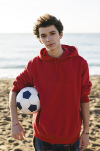 Portrait of confident teenage boy holding soccer ball while standing at beach against sea