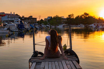 Rear view of women sitting on pier over lake against sky during sunset