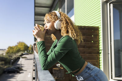 Side view of young curly haired ethnic woman listening to music while resting on balcony with cup of coffee in daytime