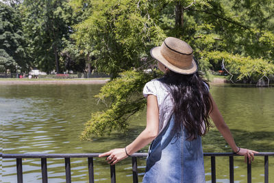 Woman wearing hat standing by lake against trees