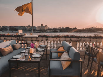 Chairs and tables at restaurant by river against sky during sunset