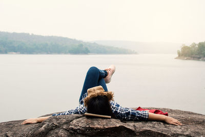 Young woman lying on rock by lake against clear sky