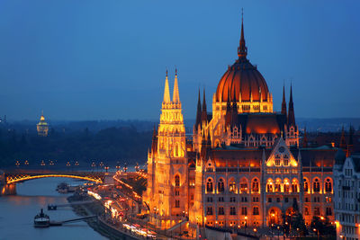 Bridge over river by illuminated hungarian parliament building against sky in city at dusk
