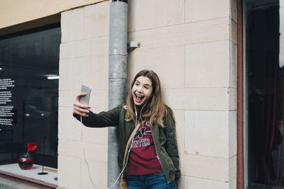 Happy girl with open mouth taking selfie through smart phone while standing against wall