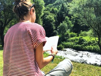 Rear view of woman reading book while sitting against river