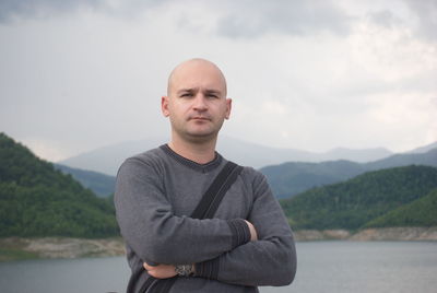 Portrait of bald man with arms crossed standing by lake against mountain