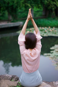 Rear view of woman with arms raised and hands clasped meditating by lake