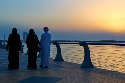 Rear view of people walking on promenade by sea against sky during sunset