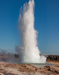 People looking at geyser against clear blue sky