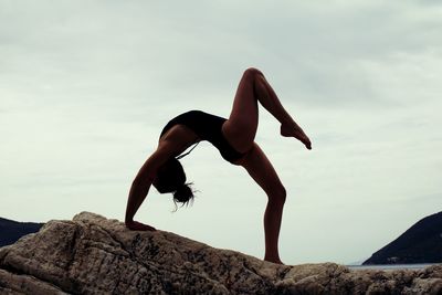 Rear view of woman in swimsuit doing yoga on rock by sea against sky