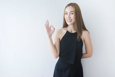 Portrait of smiling young woman showing ok sign while standing against wall