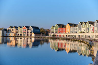 Reflection of buildings in lake against clear blue sky