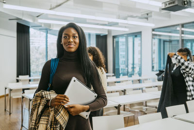 Portrait of smiling young woman holding jacket and laptop in classroom