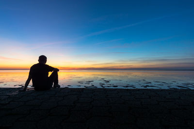 Silhouette man sitting on beach against sky during sunset