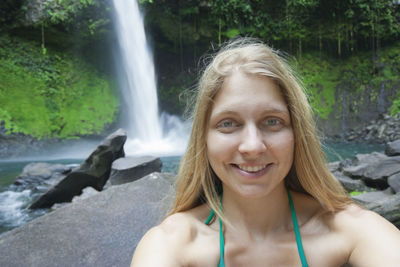 Close-up portrait of woman smiling while standing against waterfall in forest