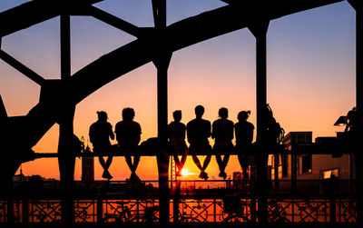 Rear view of silhouette friends sitting on hackerbrucke railway bridge against sky during sunset