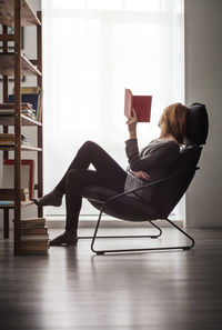 Woman holding book and relaxing on chair against window at home