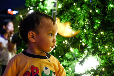 Close-up of baby boy standing by illuminated christmas tree at night
