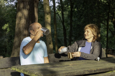 Man and woman sitting on bench rehydrating with water in a metal cup after trekking in the mountains
