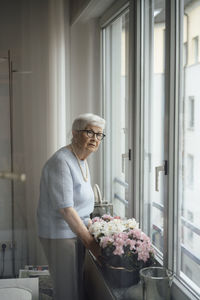 Senior woman standing by flowers and window at home