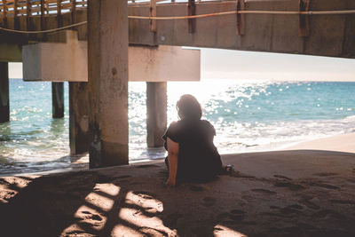 Rear view of woman sitting under pier at beach