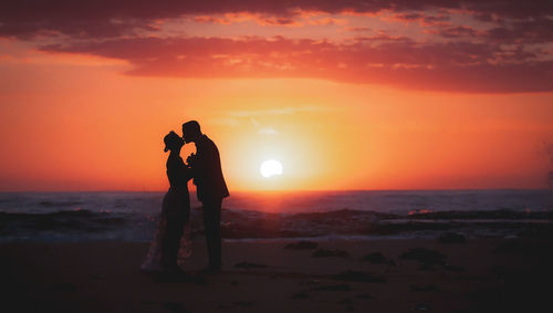Silhouette couple kissing while standing on beach against sky during sunset