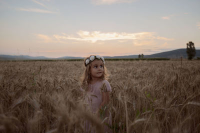 Girl standing in wheat field against sky during sunset