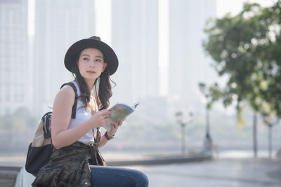 Portrait of young woman holding camera while standing outdoors