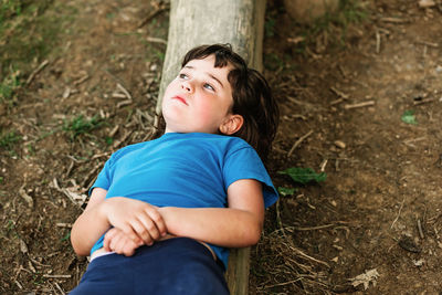 From above relaxed thoughtful girl in casual clothes lying on log near bike while spending sunny summer day in park looking away