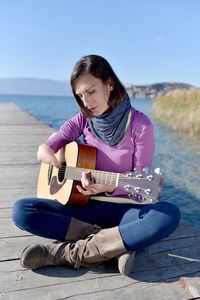 Woman playing guitar while sitting on pier by lake