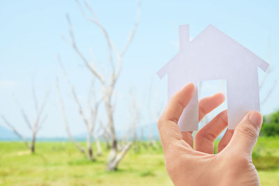 Cropped hand of person holding paper house against sky