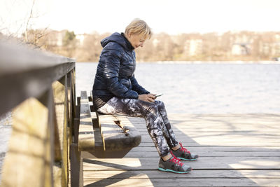 Senior woman in sportswear using phone while sitting on bench by lake