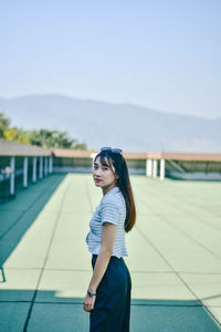 Portrait of young woman standing on floor at building terrace against sky
