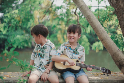 Portrait of girl playing guitar while sitting by brother on tree trunk