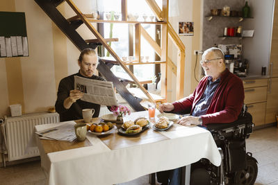 Young man showing newspaper to retired senior father with disability while eating breakfast at home
