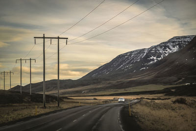 Power cable lines and road by mountain against sky during sunset