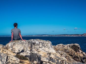 Rear view of man sitting on rock by sea against blue sky