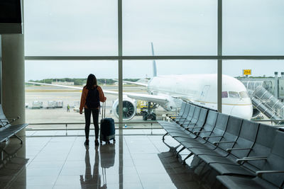 Woman with suitcase in hand, looks out the airport window while waiting for her flight.