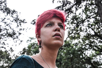 Low angle view of thoughtful young woman with dyed hair standing against sky in forest