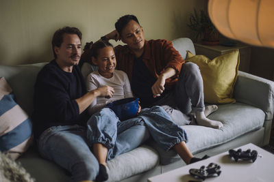 Gay couple watching tv with daughter eating popcorn while sitting on sofa at home