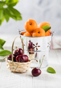 Ripe cherries in a basket and fresh apricots in a bucket on a white wooden background. 