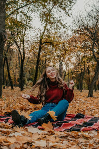 Smiling young woman sitting on autumn leaves against trees in forest