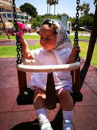 Cute baby girl holding flowers while sitting on swing in playground